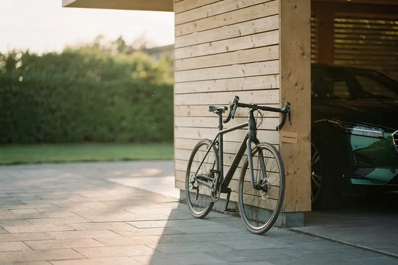 Fahrrad an der Holzwand eines Carports neben der Front eines grünen Autos im warmen Abendlicht