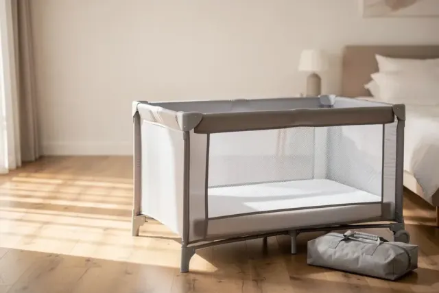 Baby-Reisebett mit Netzseiten und flacher Matratze ohne Kissen im Schlafzimmer, daneben die Tasche
